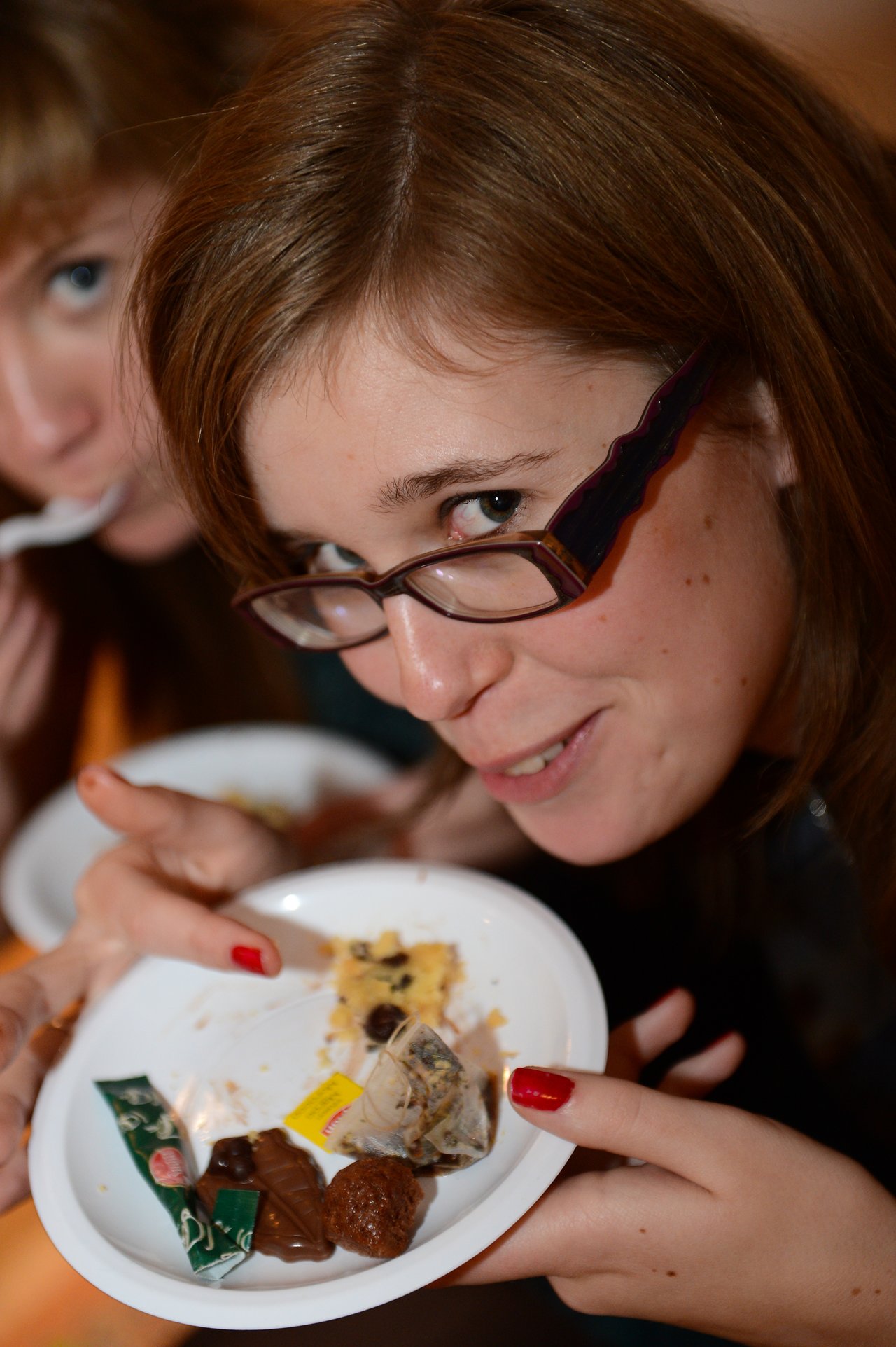 A woman with glasses smiles while holding a plate with chocolates, cake, and a tea bag at a party.