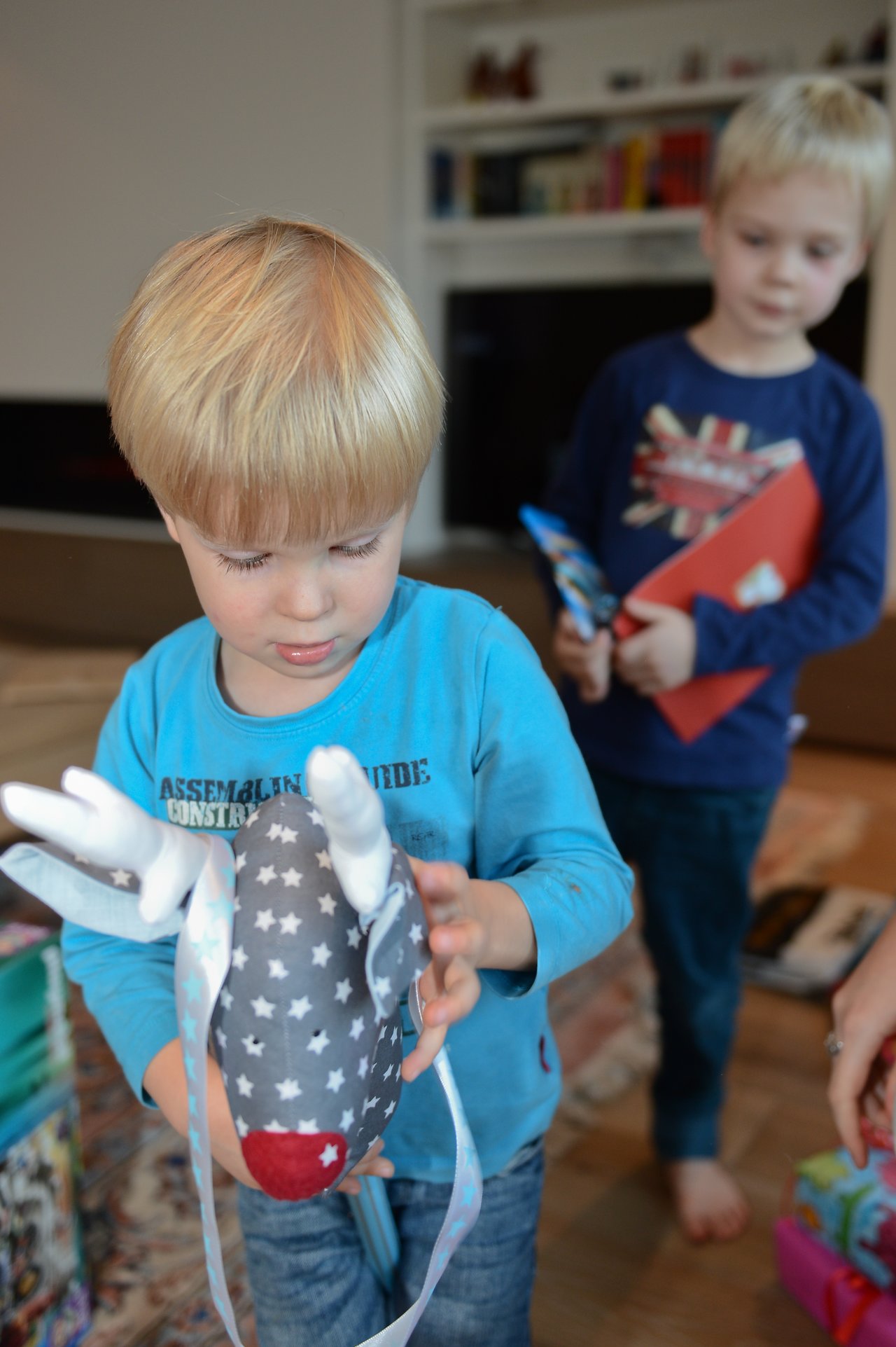 A young child in a blue shirt holds a stuffed reindeer toy, while another child stands behind him.