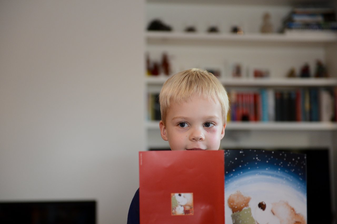 A young child holds up a festive card, partially covering their face, in a home setting.