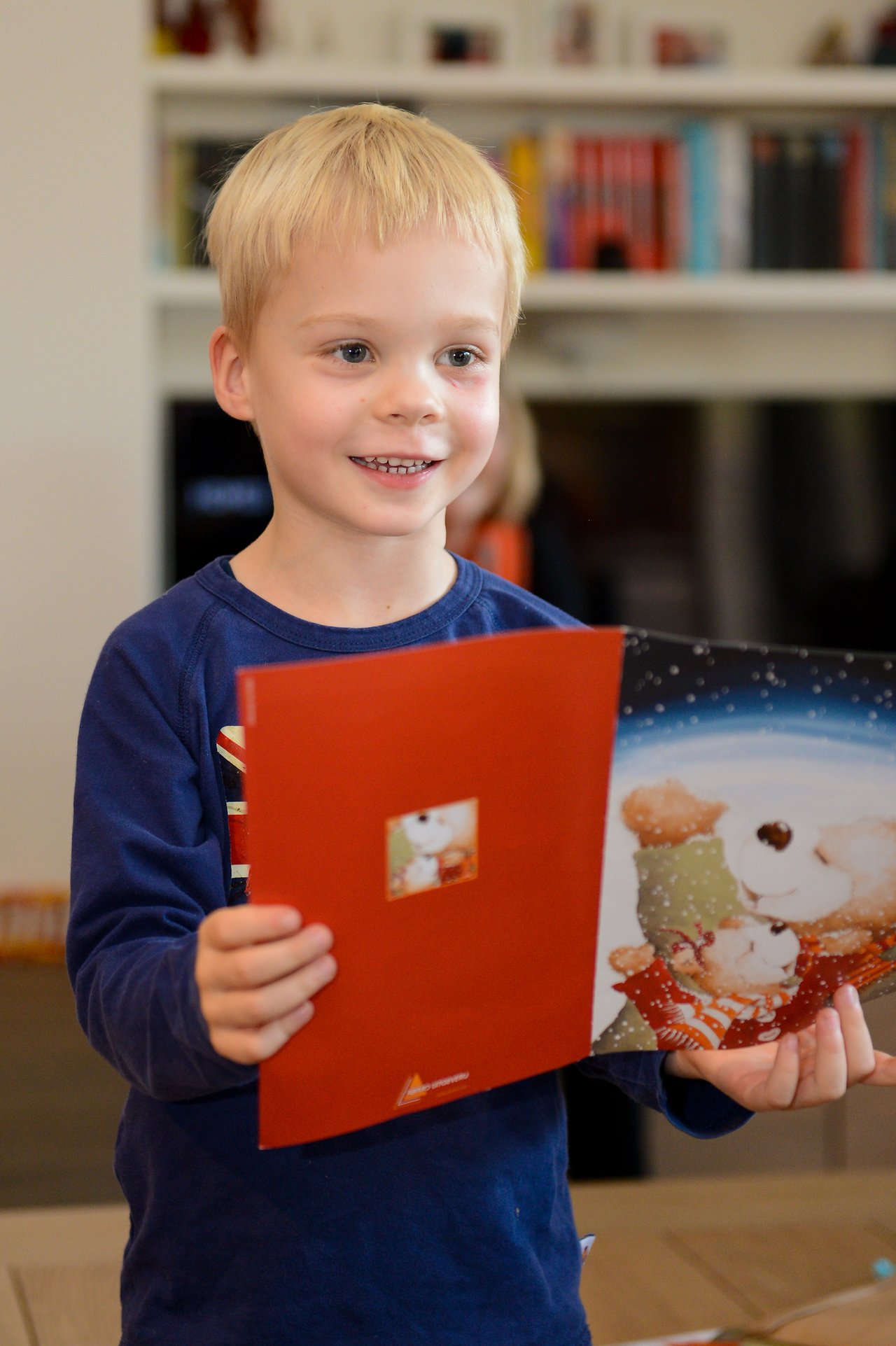 A smiling child in a blue shirt holds up a festive greeting card with a teddy bear illustration.