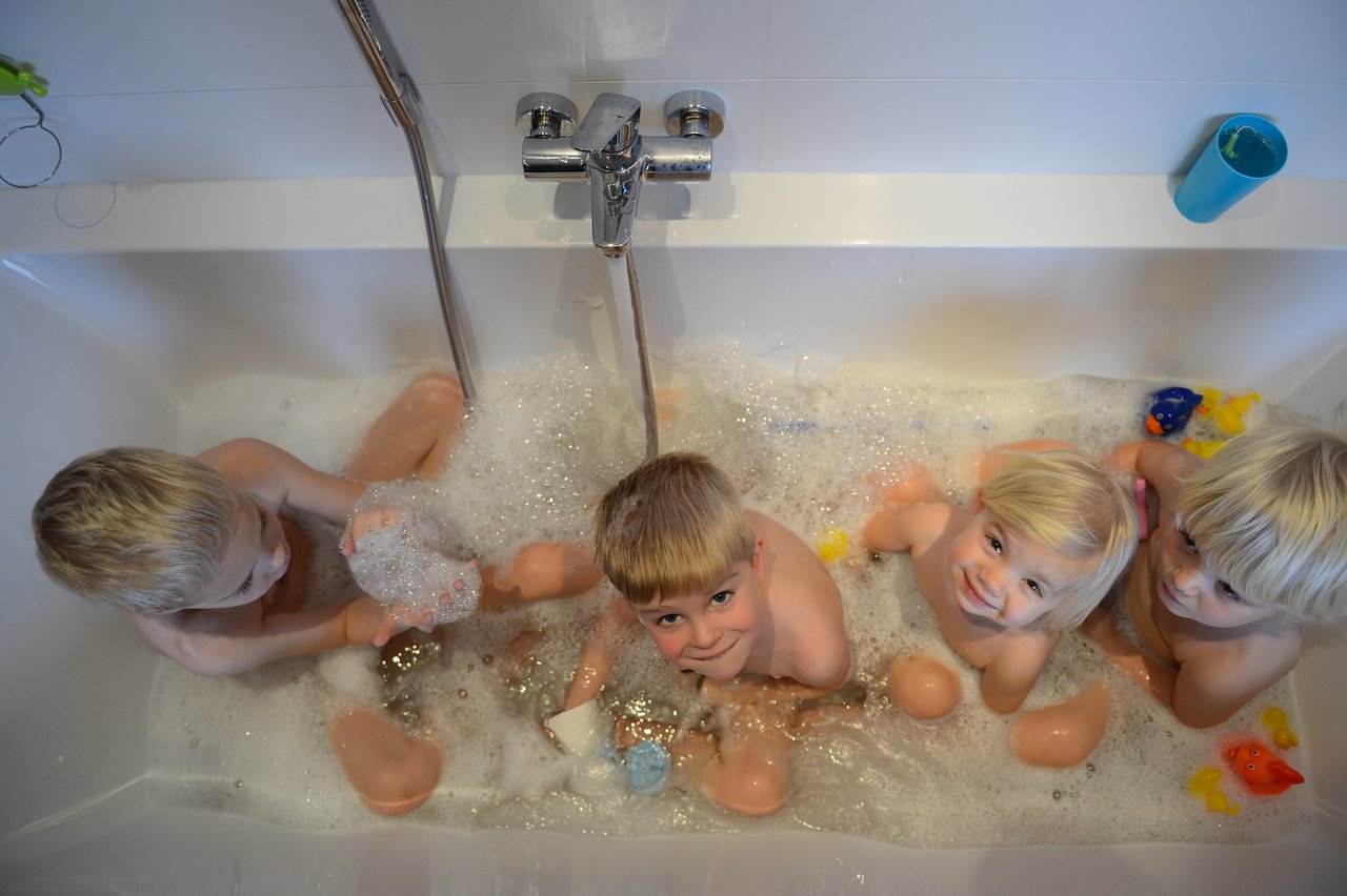 Four young children sit in a bathtub filled with bubbles, playing with bath toys and smiling.