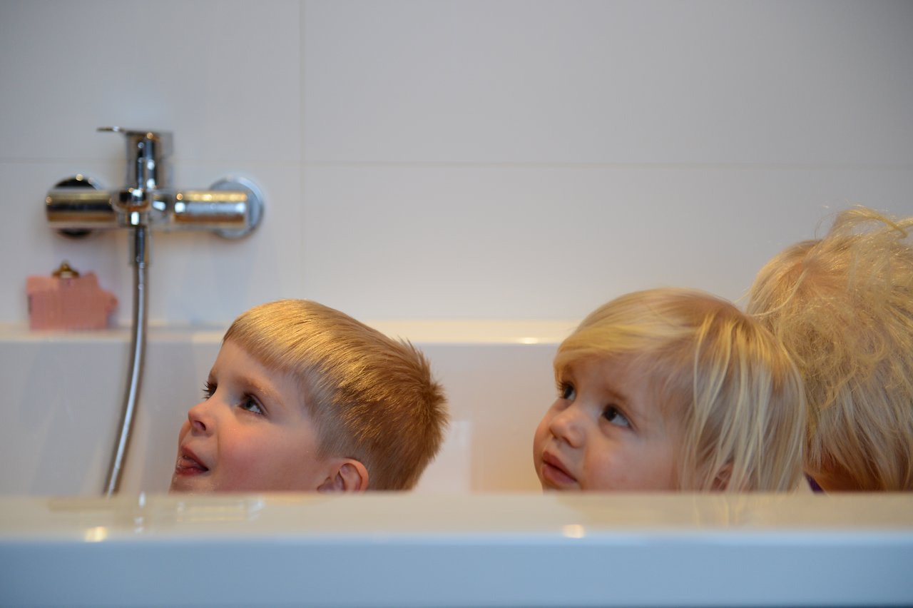 Three young children sit in a bathtub, looking up with curious expressions.