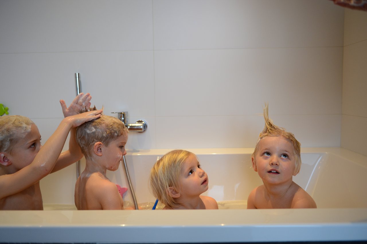 Four young children are in a bathtub, playing with shampoo bubbles and styling their hair into fun shapes.