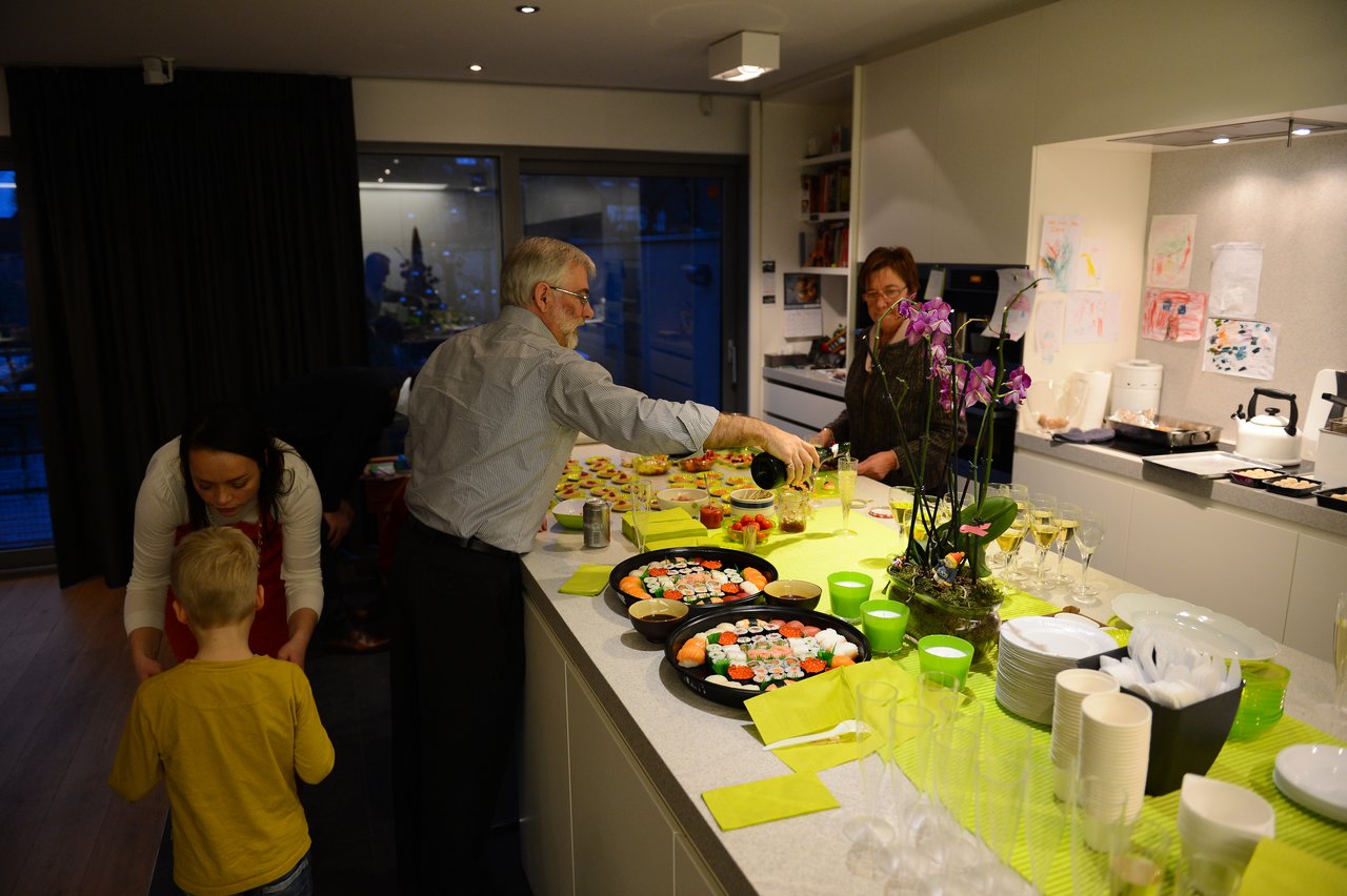 A man pours a drink at a kitchen counter with sushi and snacks, while others prepare and interact nearby.
