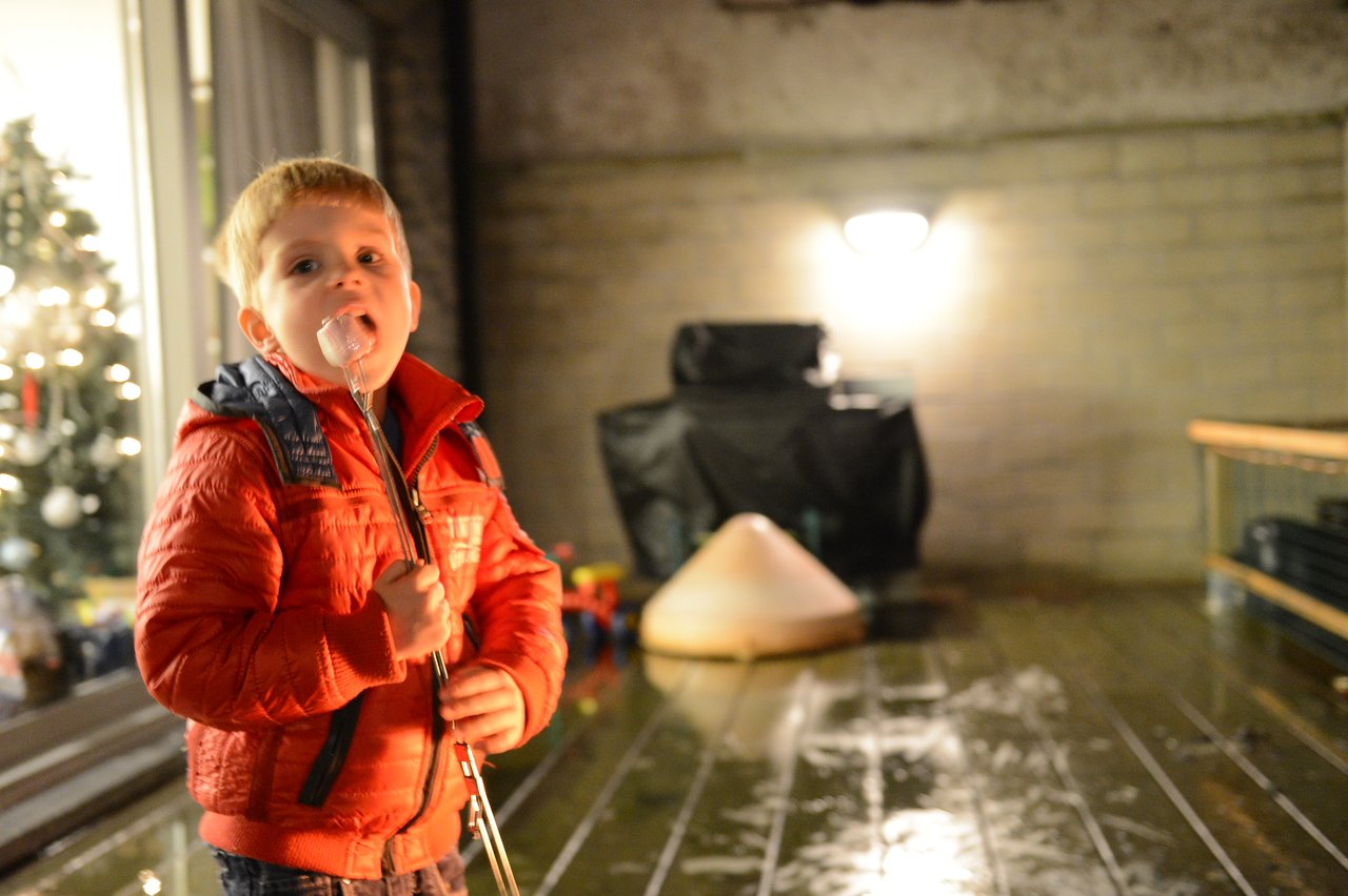 A young child in a red jacket holds a skewer with a marshmallow near their mouth, standing outside.