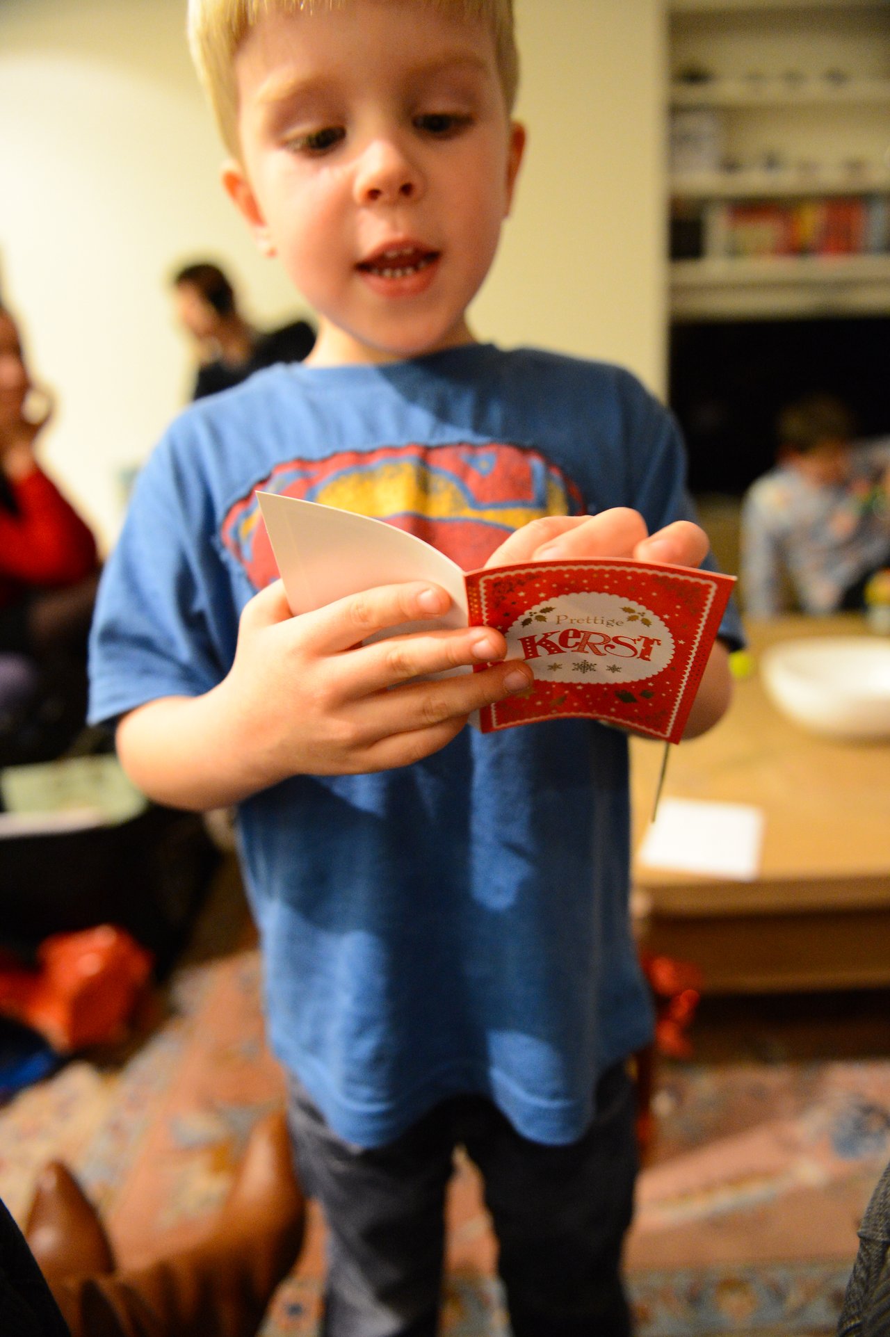 A young child in a blue Superman shirt reads a festive card aloud in a warmly lit room.