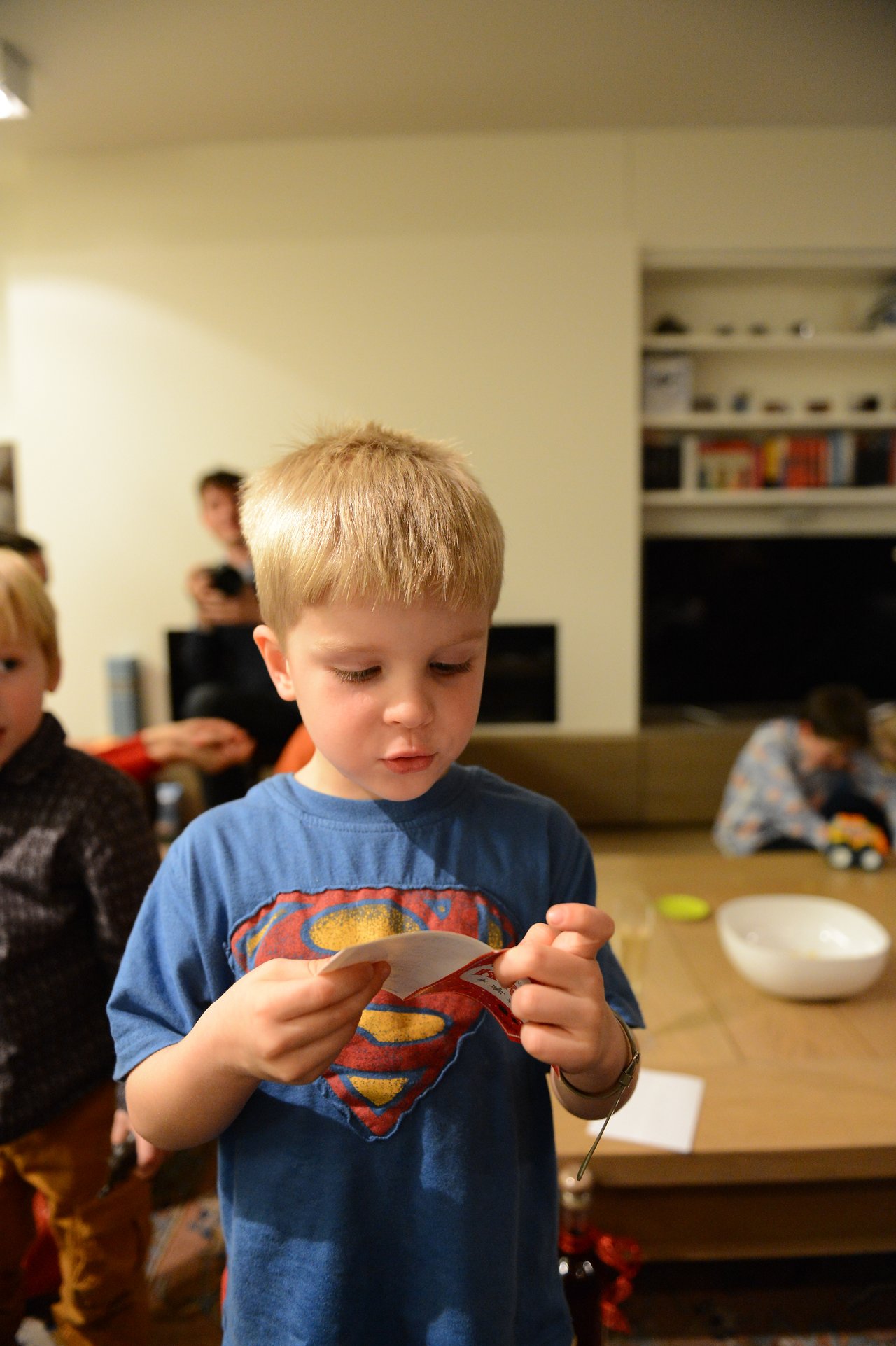 A young boy in a blue Superman shirt reads a small card while standing in a living room.