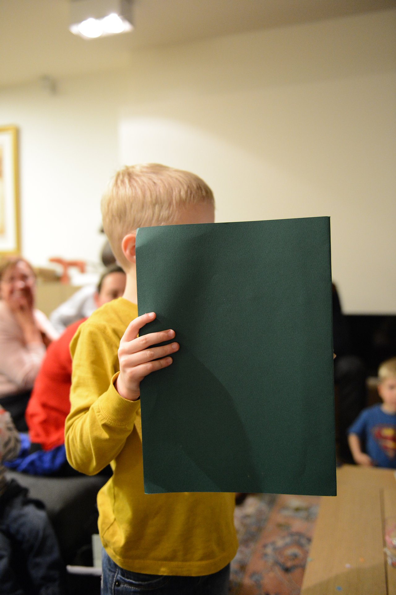 A child in a yellow shirt holds up a large green sheet of paper, partially covering their face.