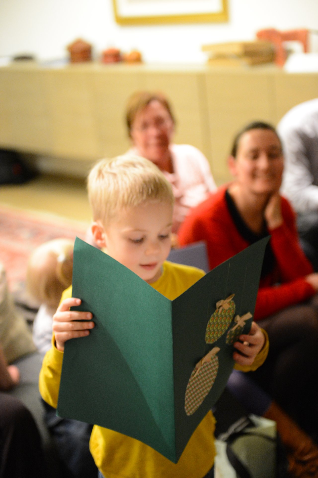 A young child in a yellow shirt holds a large green card with decorations, while adults watch in the background.