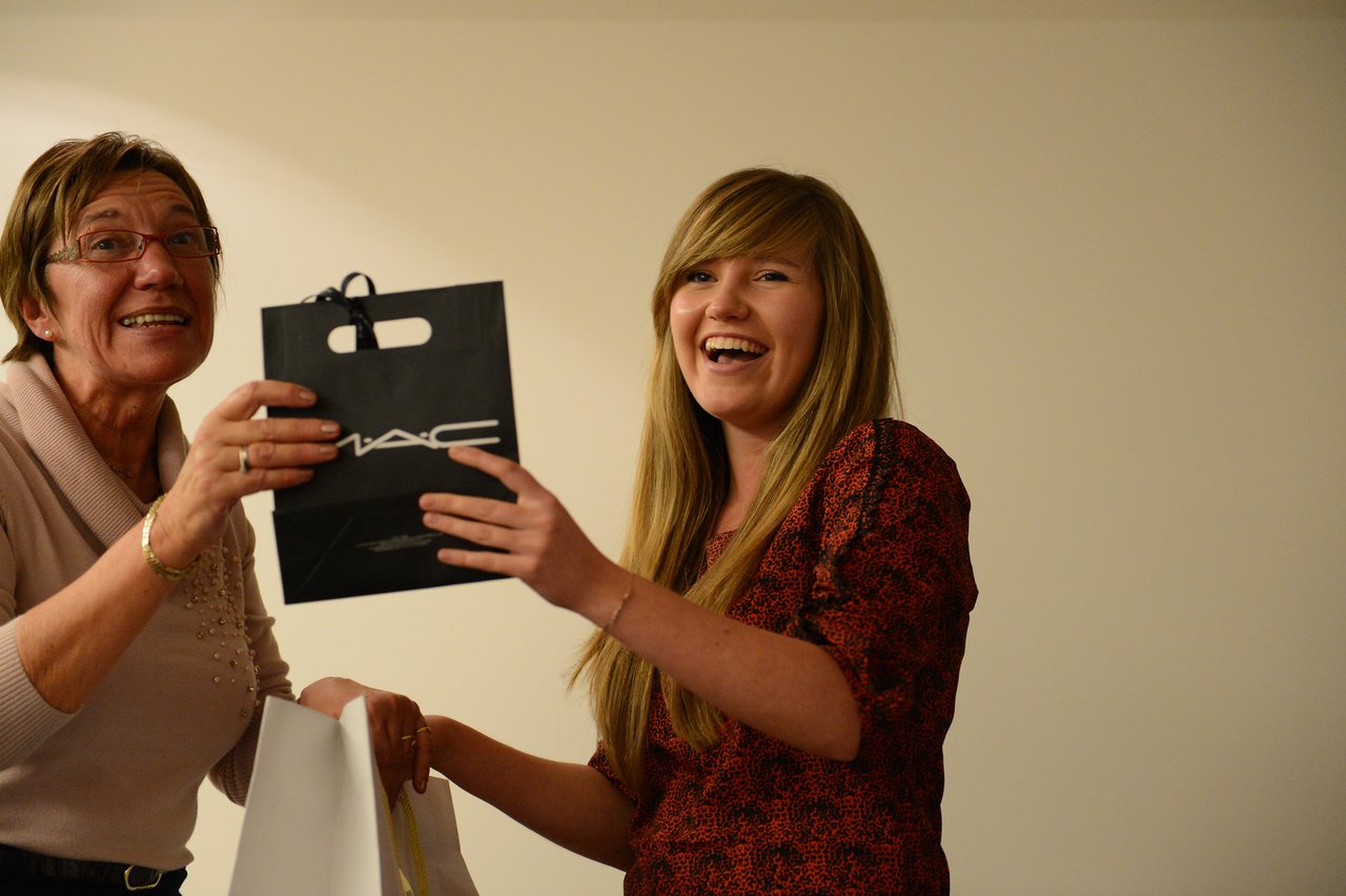 Two women smiling as one hands a black MAC gift bag to the other.