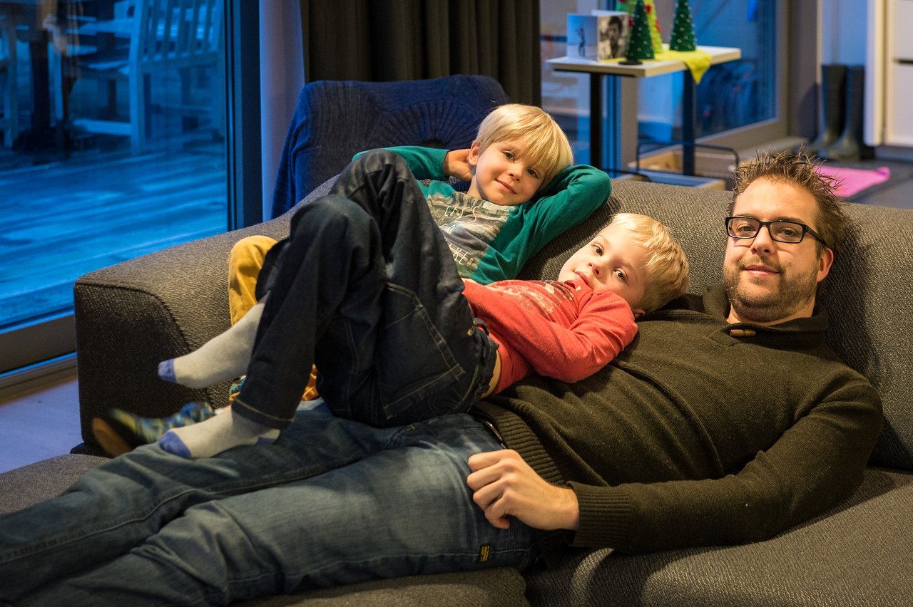 A man and two children relax on a couch, smiling and looking at the camera in a cozy indoor setting.