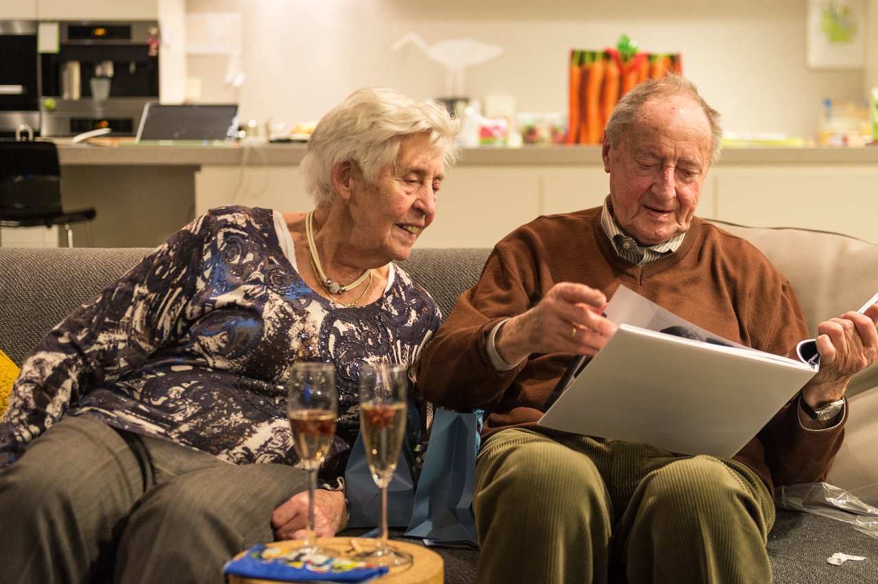 An elderly couple sits on a couch, looking through a photo album with two glasses of champagne nearby.