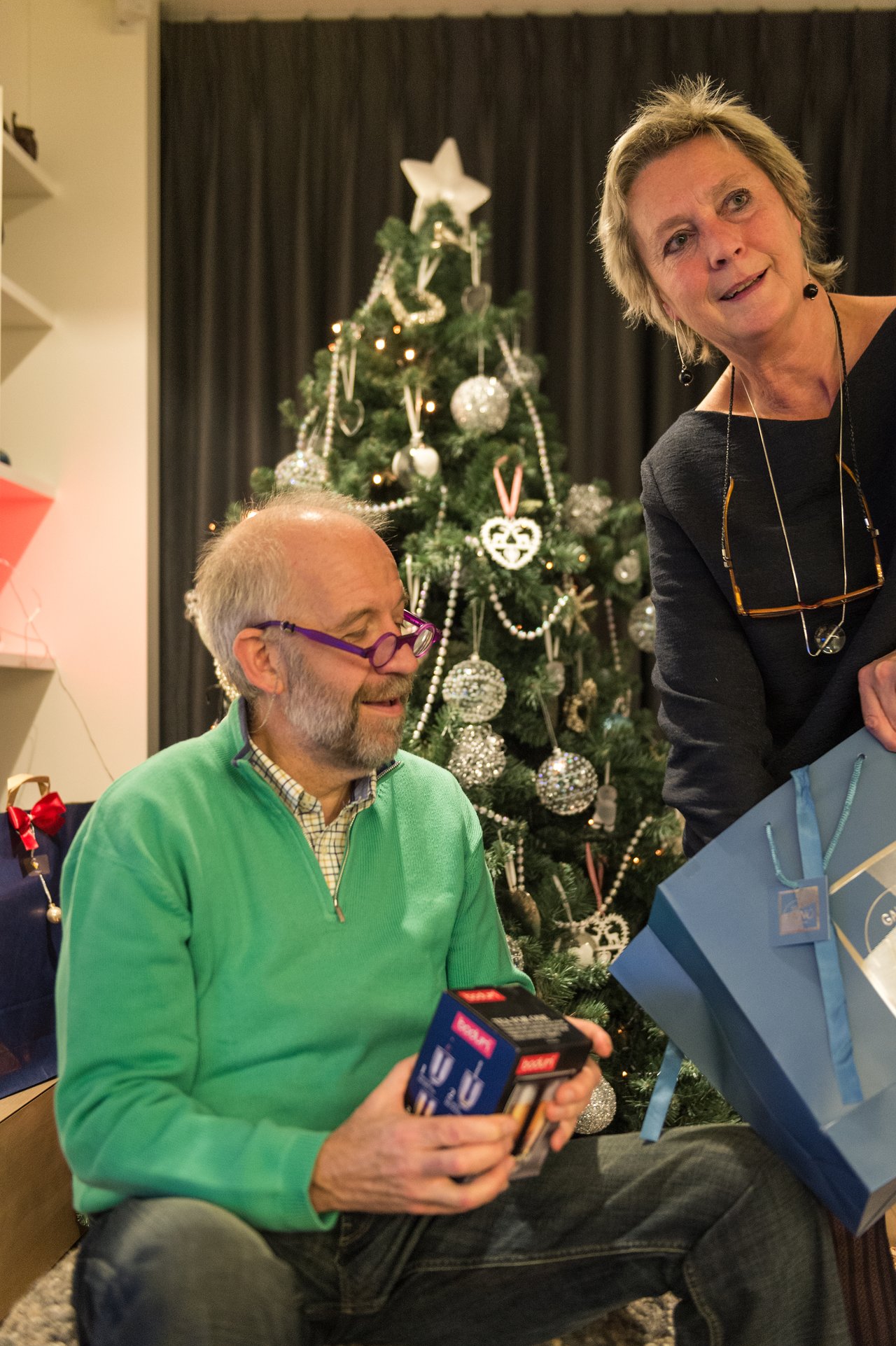 A man in a green sweater holds a gift while a woman hands him a blue shopping bag near a Christmas tree.