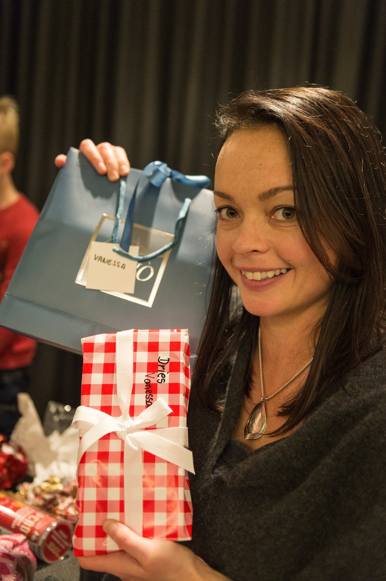 A woman smiles while holding a gift bag and a wrapped present during a New Year's celebration.