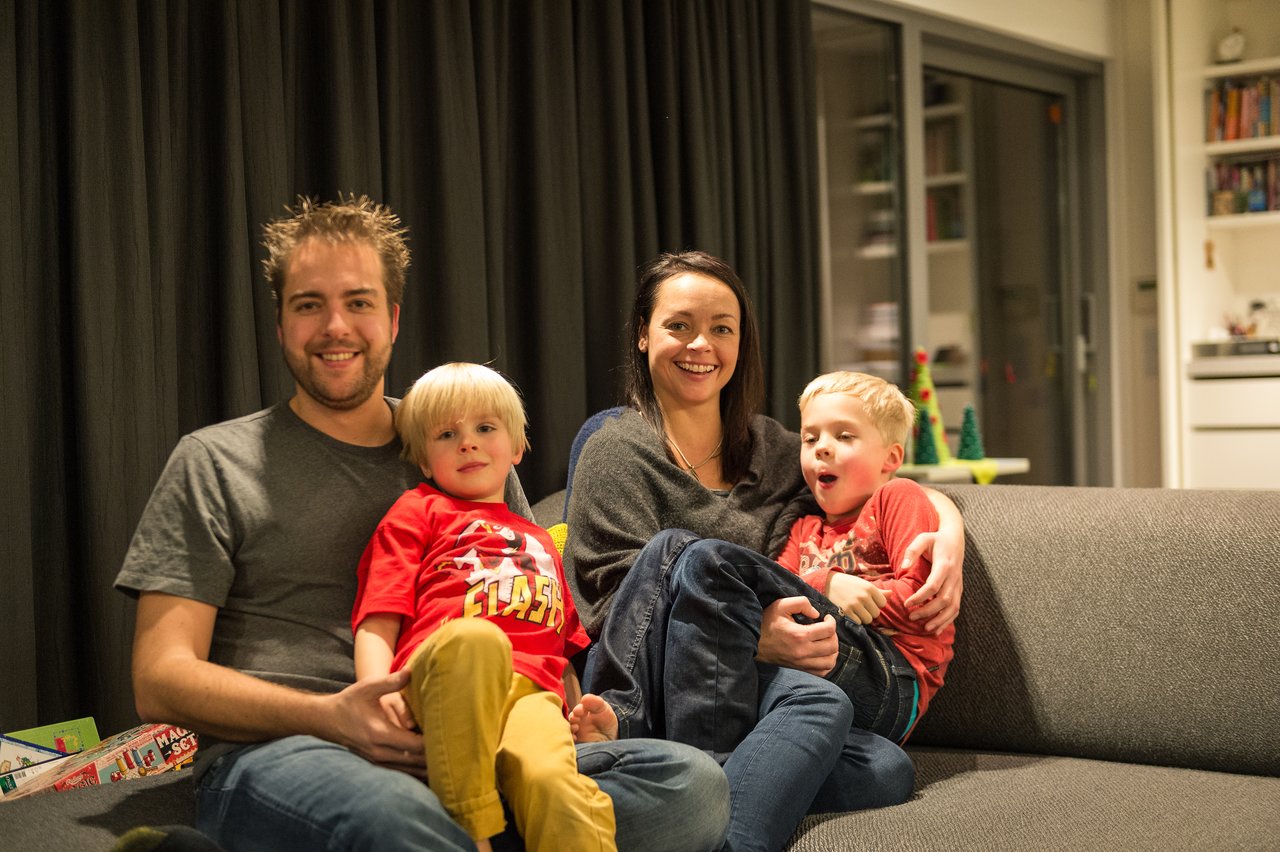A smiling family of four sits together on a couch, with two young children on their parents' laps.