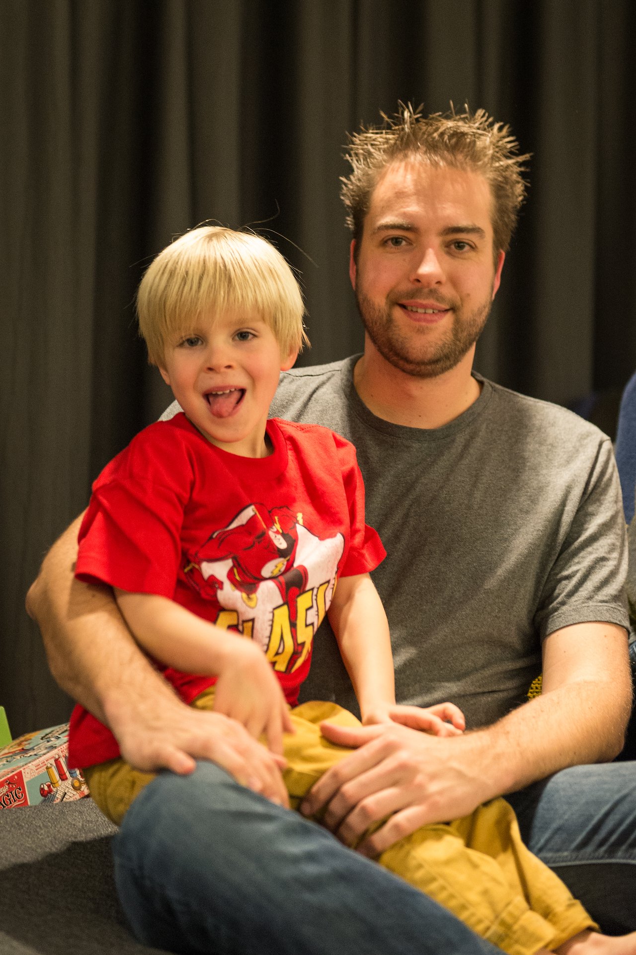 A man sits on a couch, smiling, while a young child in a red shirt sits on his lap, playful.