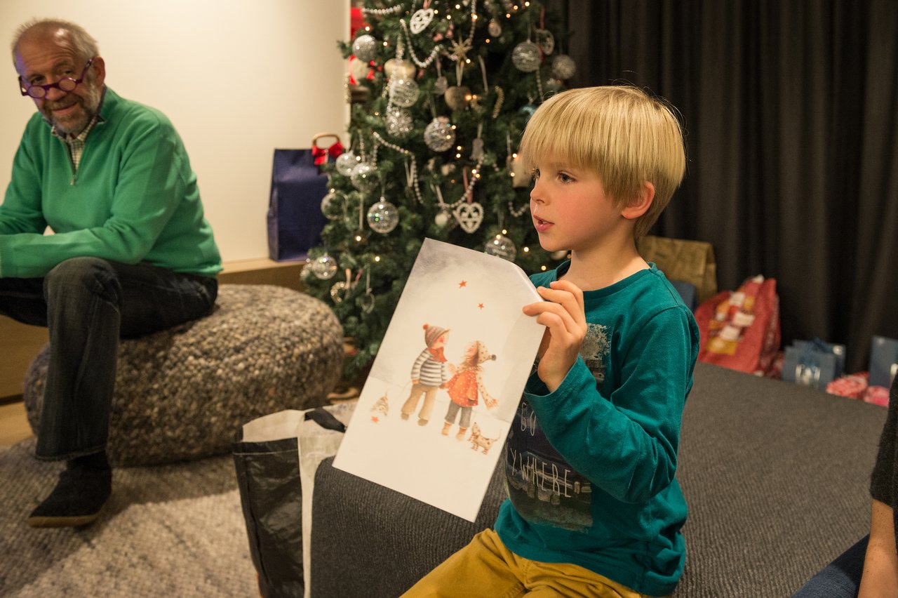 A young boy holds up a festive drawing while sitting near a decorated Christmas tree with presents underneath.