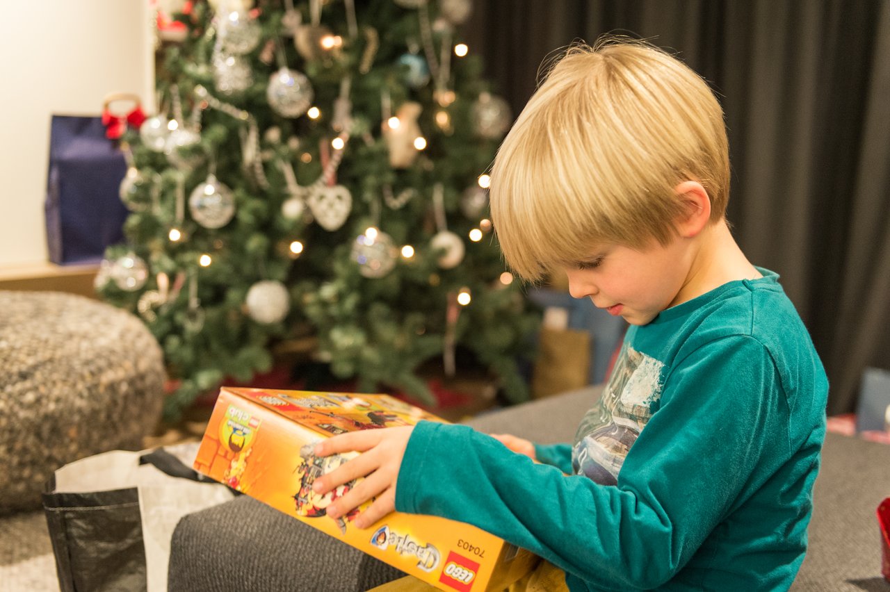 A young boy in a green shirt excitedly opens a LEGO box near a decorated Christmas tree.