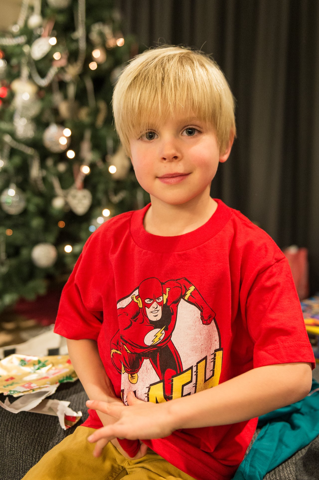 A young child in a red Flash t-shirt sits in front of a decorated Christmas tree with presents nearby.
