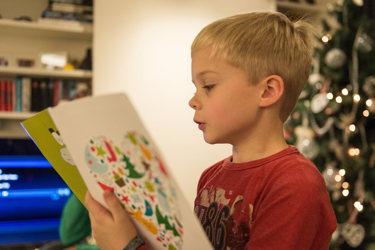 A young boy in a red shirt reads from a festive book near a decorated Christmas tree.
