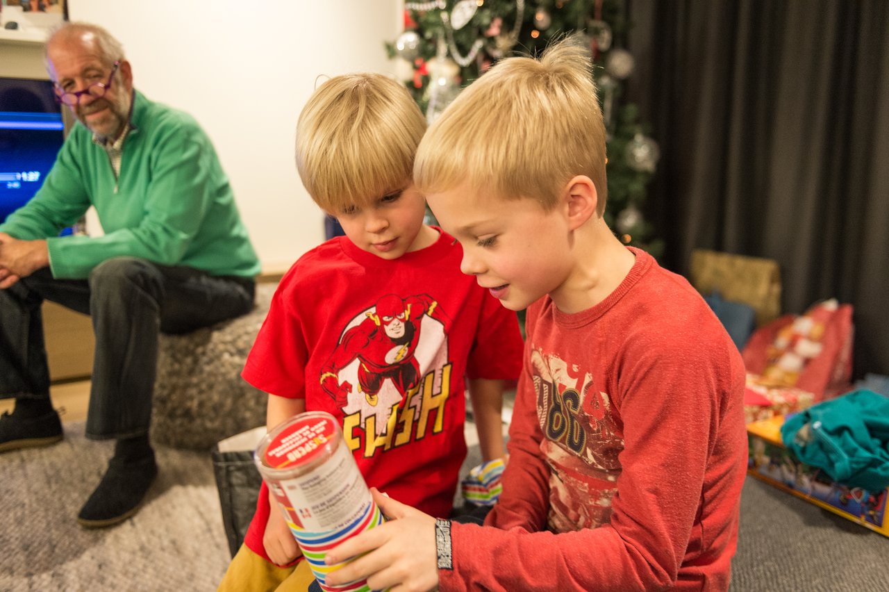 Two children excitedly examine a gift while an older man watches from the background near a decorated tree.