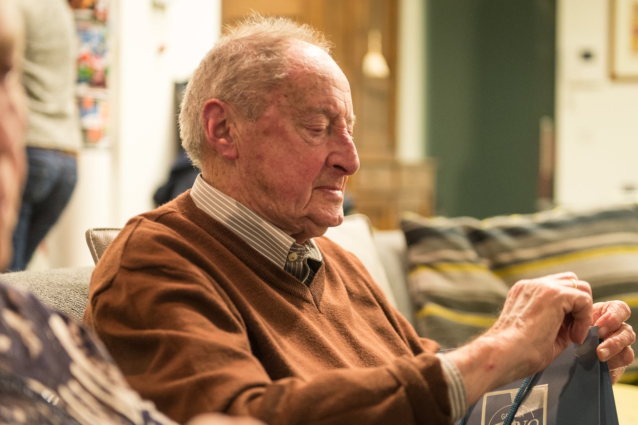 An elderly man sits on a couch, opening a gift bag with a focused expression.