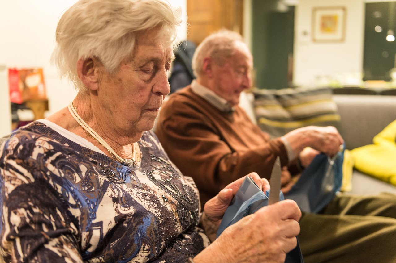Elderly woman and man sitting indoors, cutting blue plastic with knives while focusing on their task.