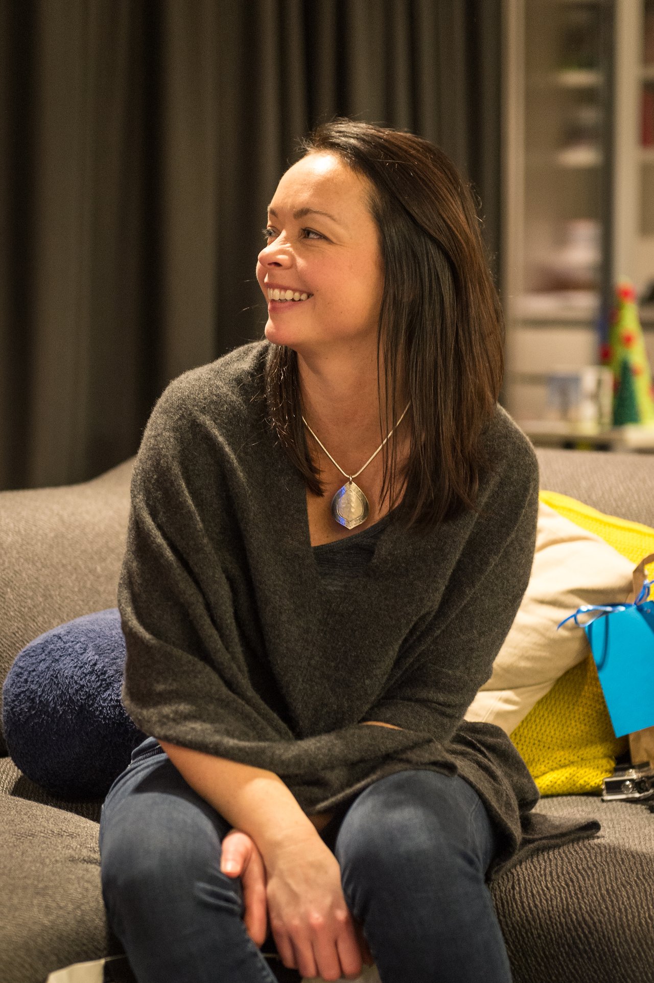 A woman sits on a couch, smiling and looking to the side, wearing a dark sweater and necklace.