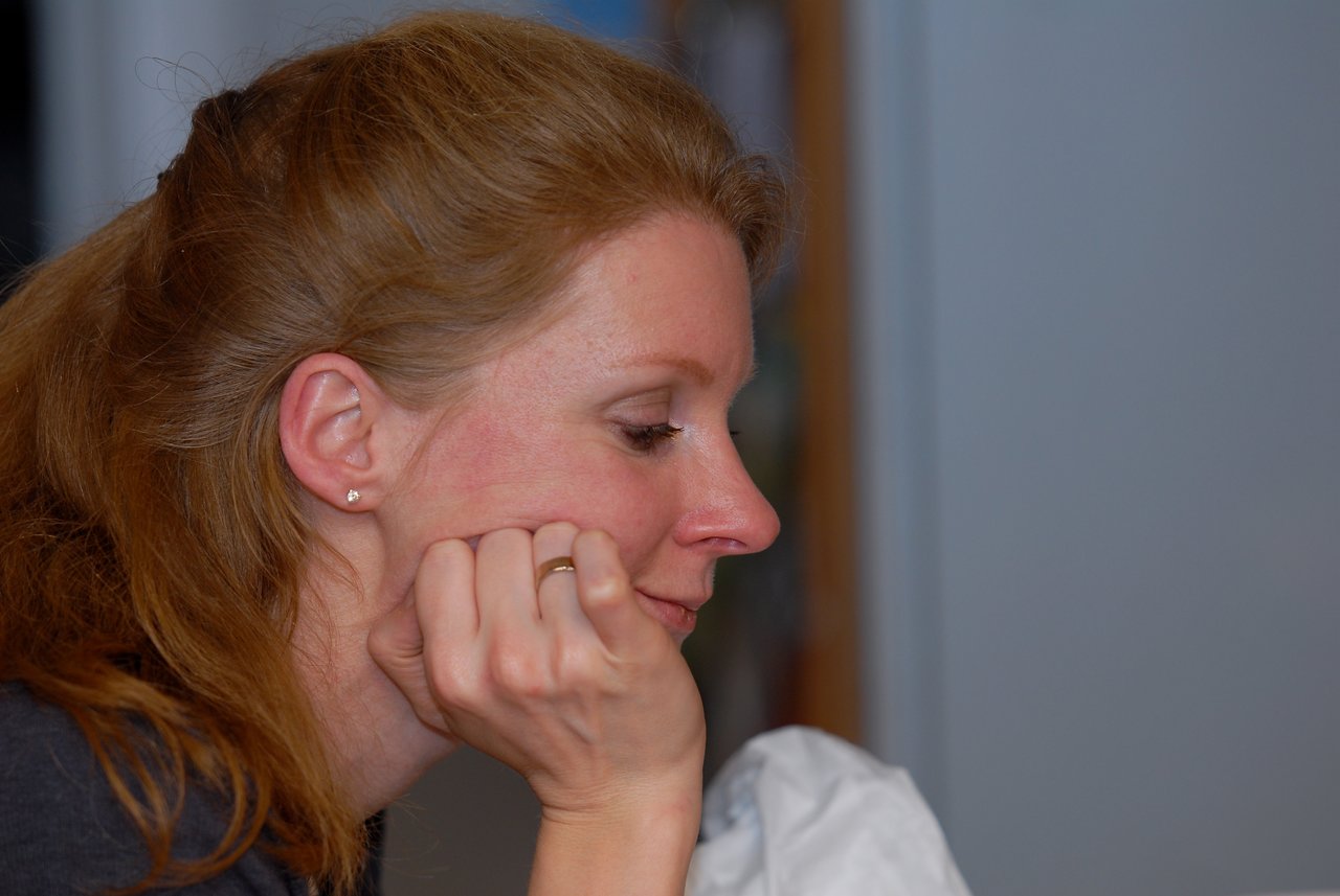 A woman with red hair rests her chin on her hand, looking downward with a neutral expression.