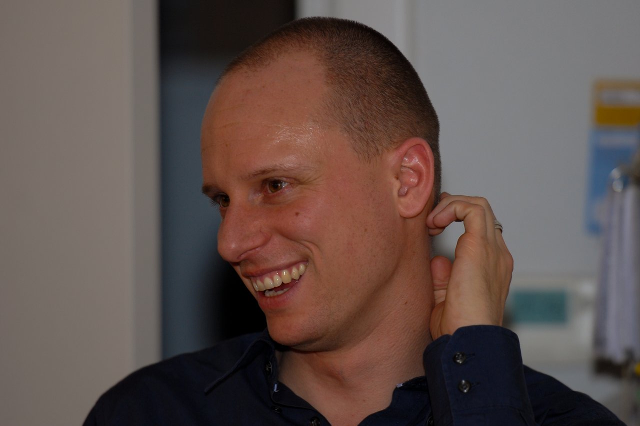 A man in a dark shirt smiles while touching his ear at a New Year's Eve party.