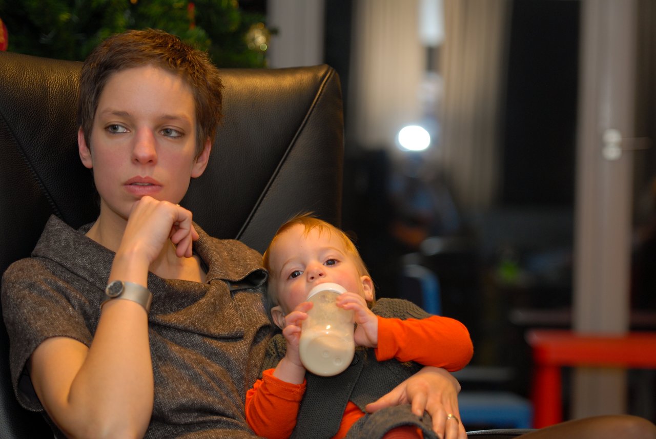 A woman sits in a chair, looking away, while a baby drinks from a bottle in her arms.