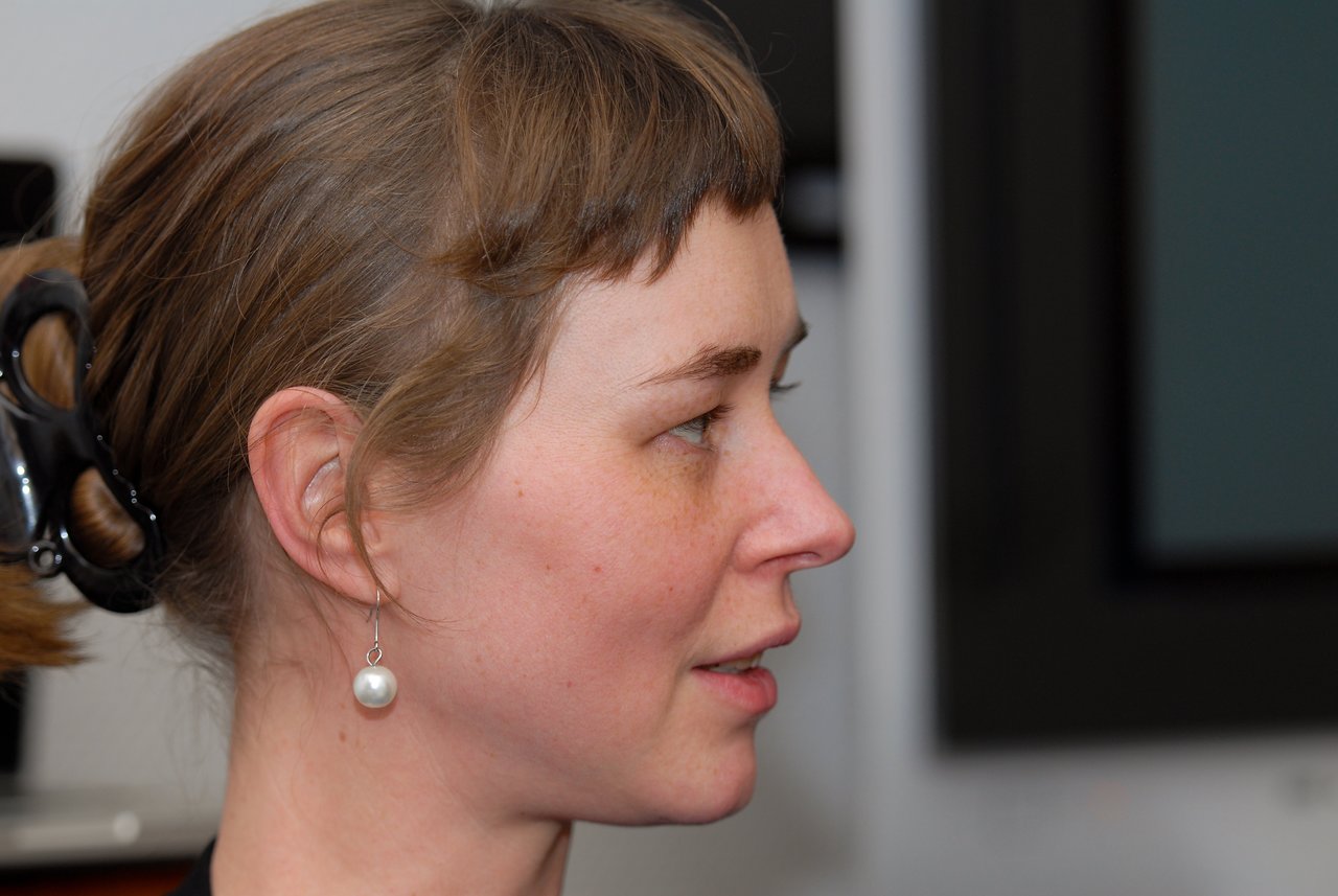 A woman with light brown hair and a pearl earring looks to the side at an indoor gathering.
