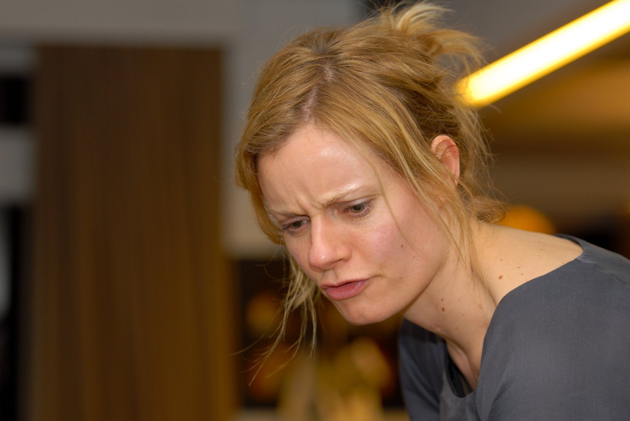 A woman with blonde hair and a gray top looks down with a focused or concerned expression at a party.