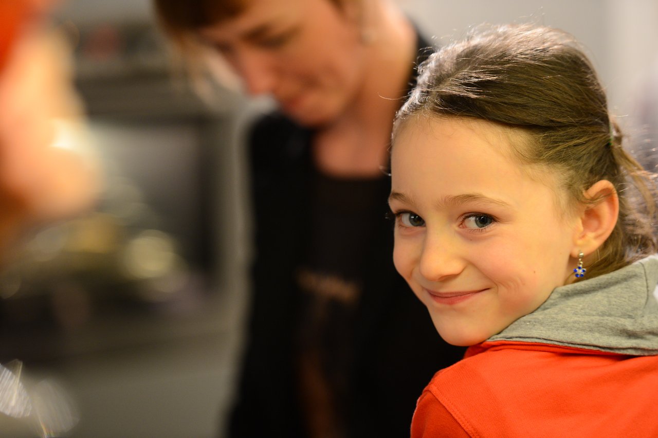 A young girl in a red hoodie smiles at the camera while a blurred adult figure is in the background.