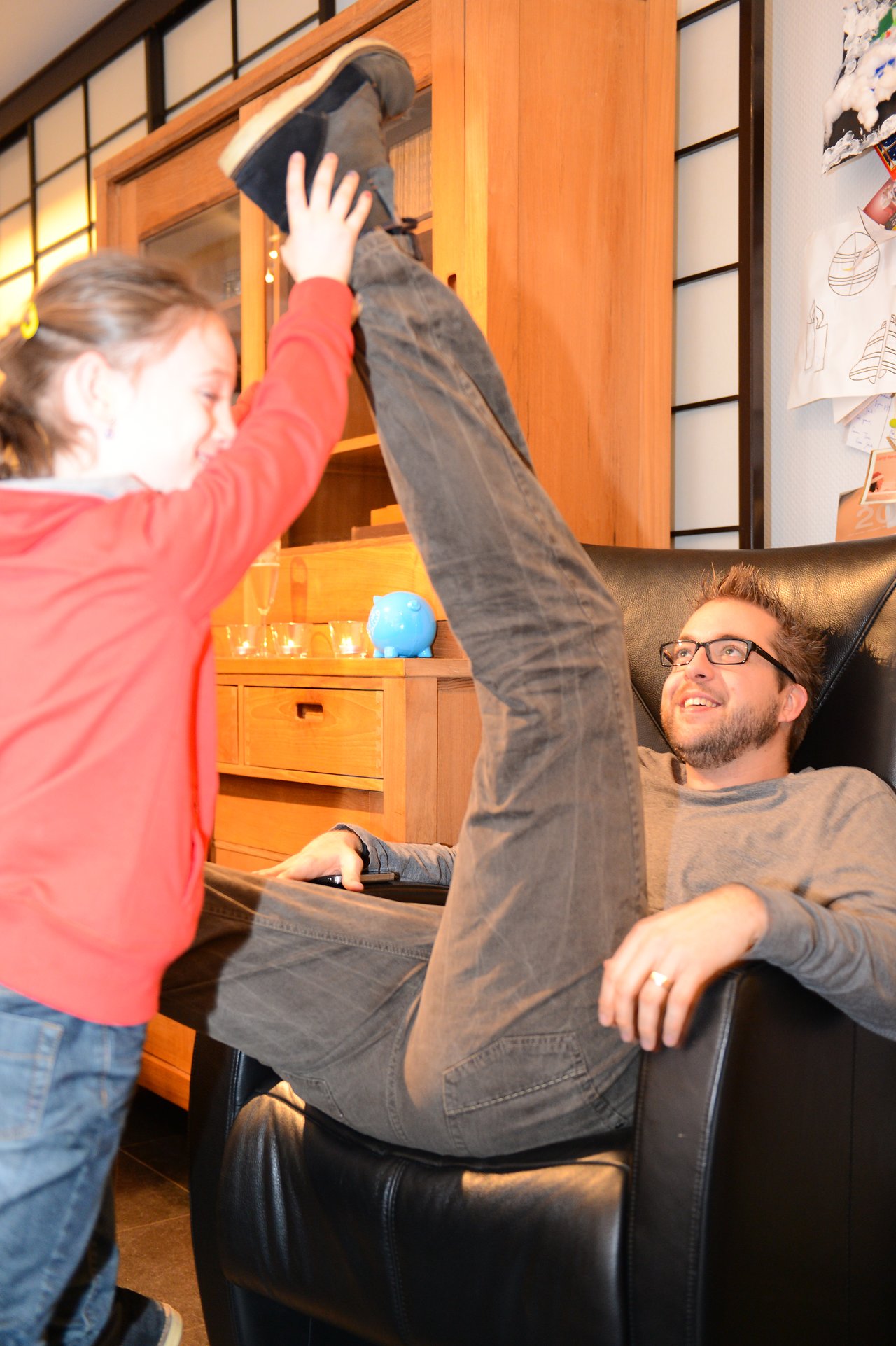 A child in a red hoodie playfully lifts an adult's leg while they sit in a black chair, smiling.