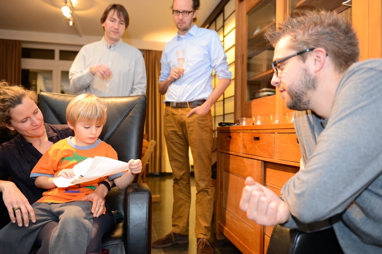 A child sits on a woman's lap, reading a paper, while three adults stand nearby holding drinks.