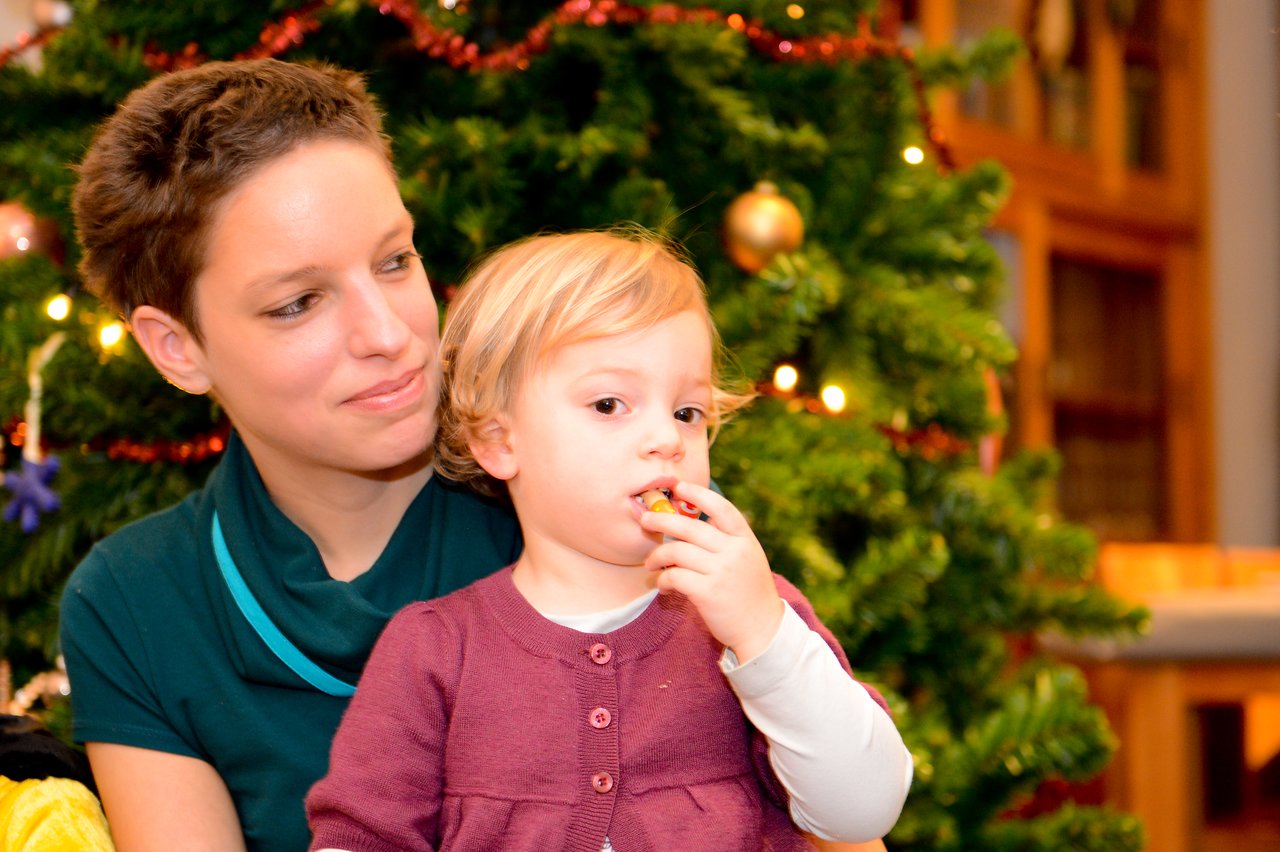 A woman smiles while holding a young child who is eating a snack near a decorated Christmas tree.