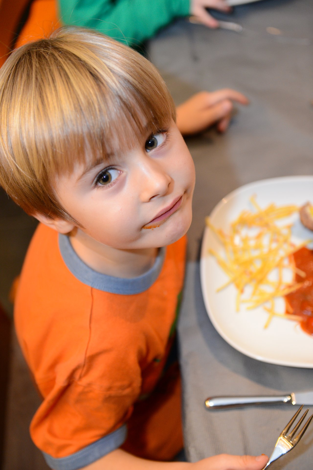 A young child in an orange shirt looks up while sitting at a table with a plate of food.