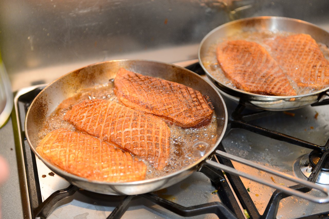 Several pieces of meat with a crisscross pattern are frying in two pans on a stovetop.
