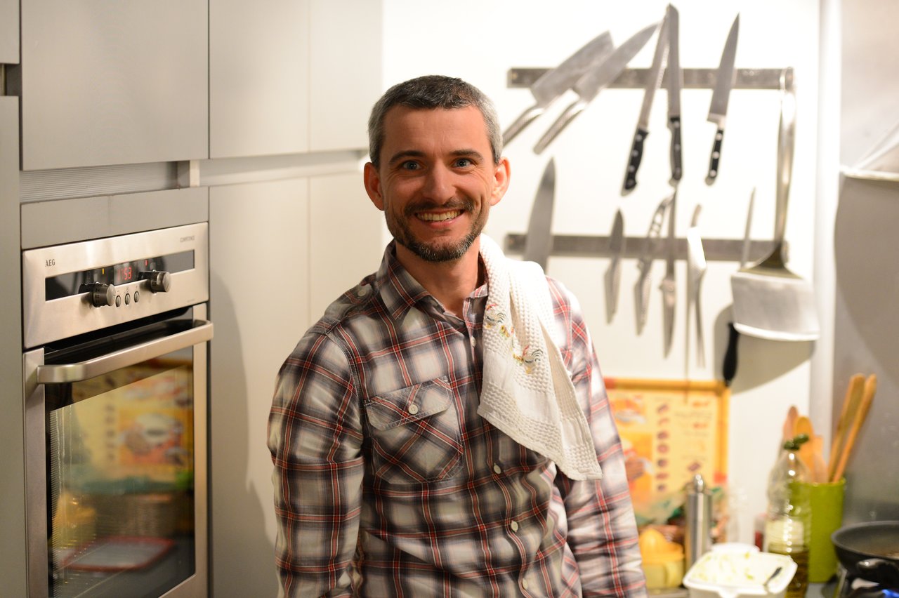 A smiling man in a plaid shirt stands in a kitchen with a towel over his shoulder.