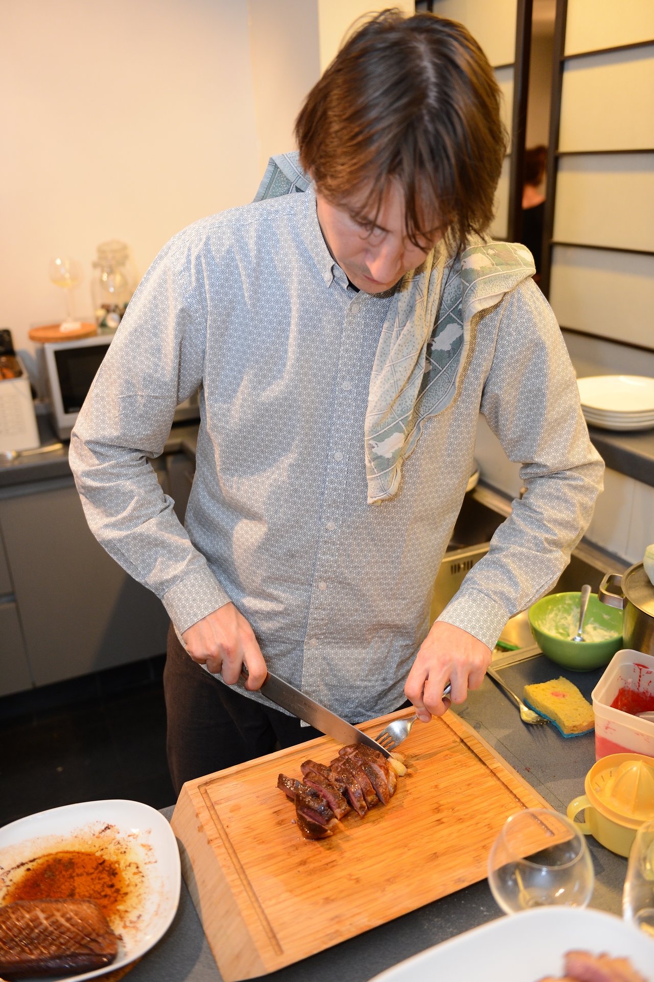 A person in a button-up shirt slices cooked meat on a wooden cutting board at a party.