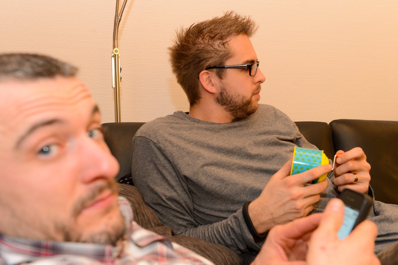 Two men sitting on a couch; one eating from a small container, the other using a phone.