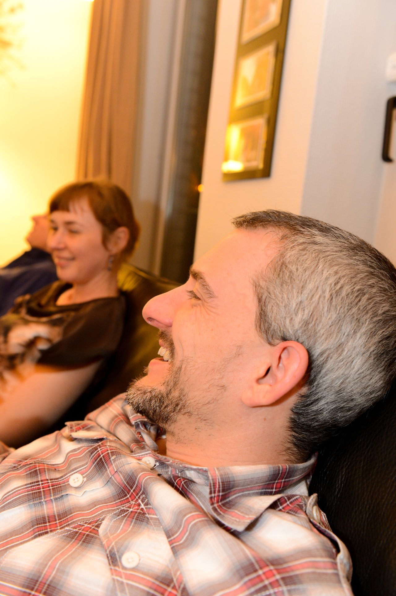 A man in a plaid shirt smiles while relaxing on a couch, with others enjoying the party in the background.