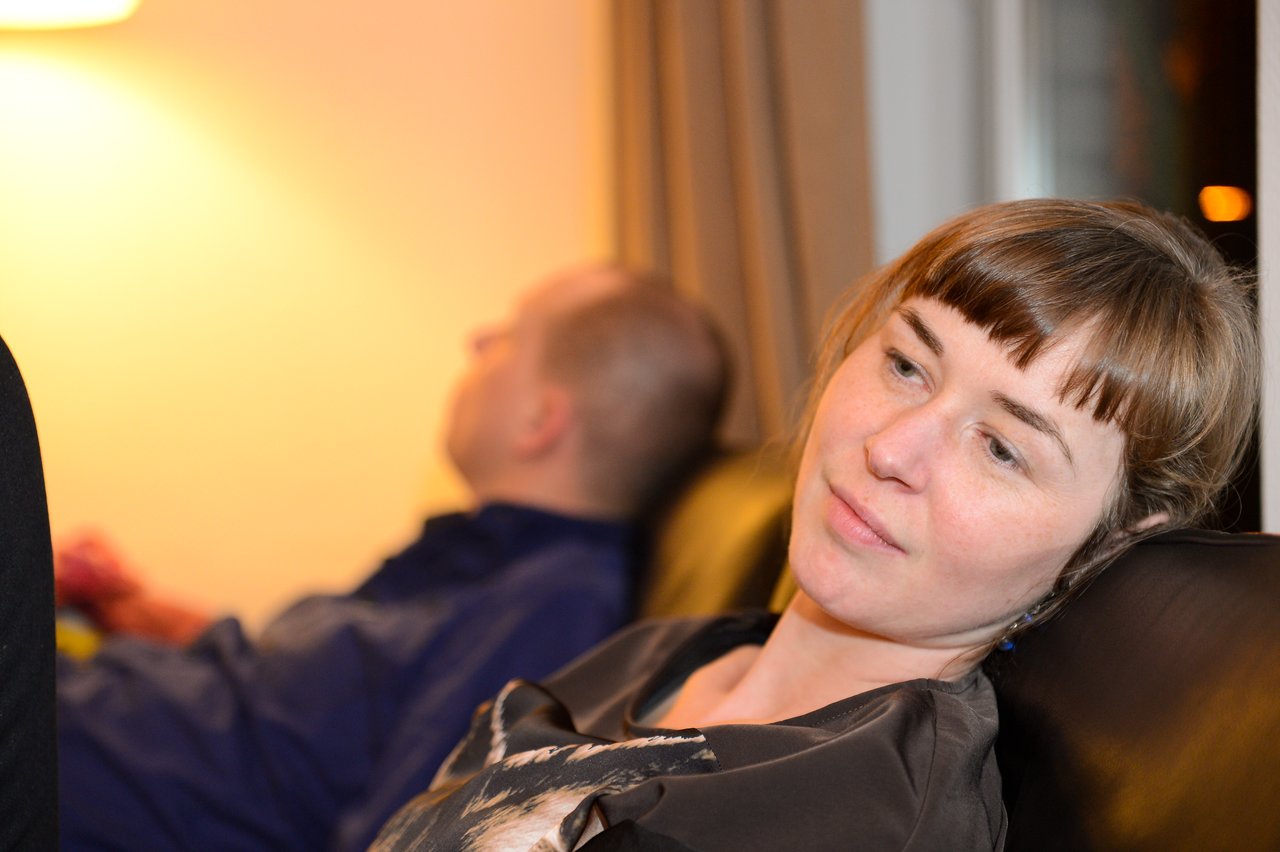 A woman leans back on a couch, looking relaxed, while a man in the background appears to be resting.