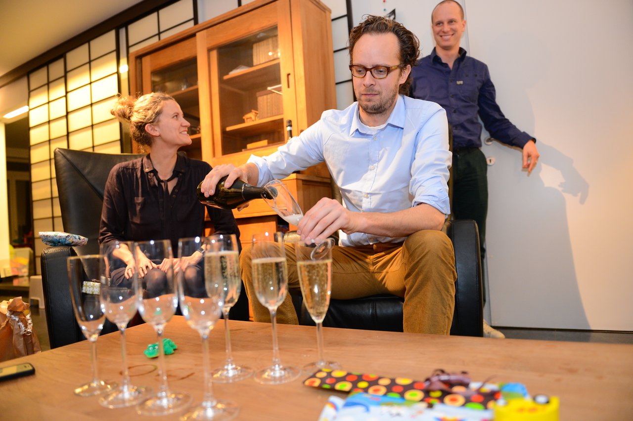 A man pours champagne into glasses while two others smile and watch during a New Year's Eve party.