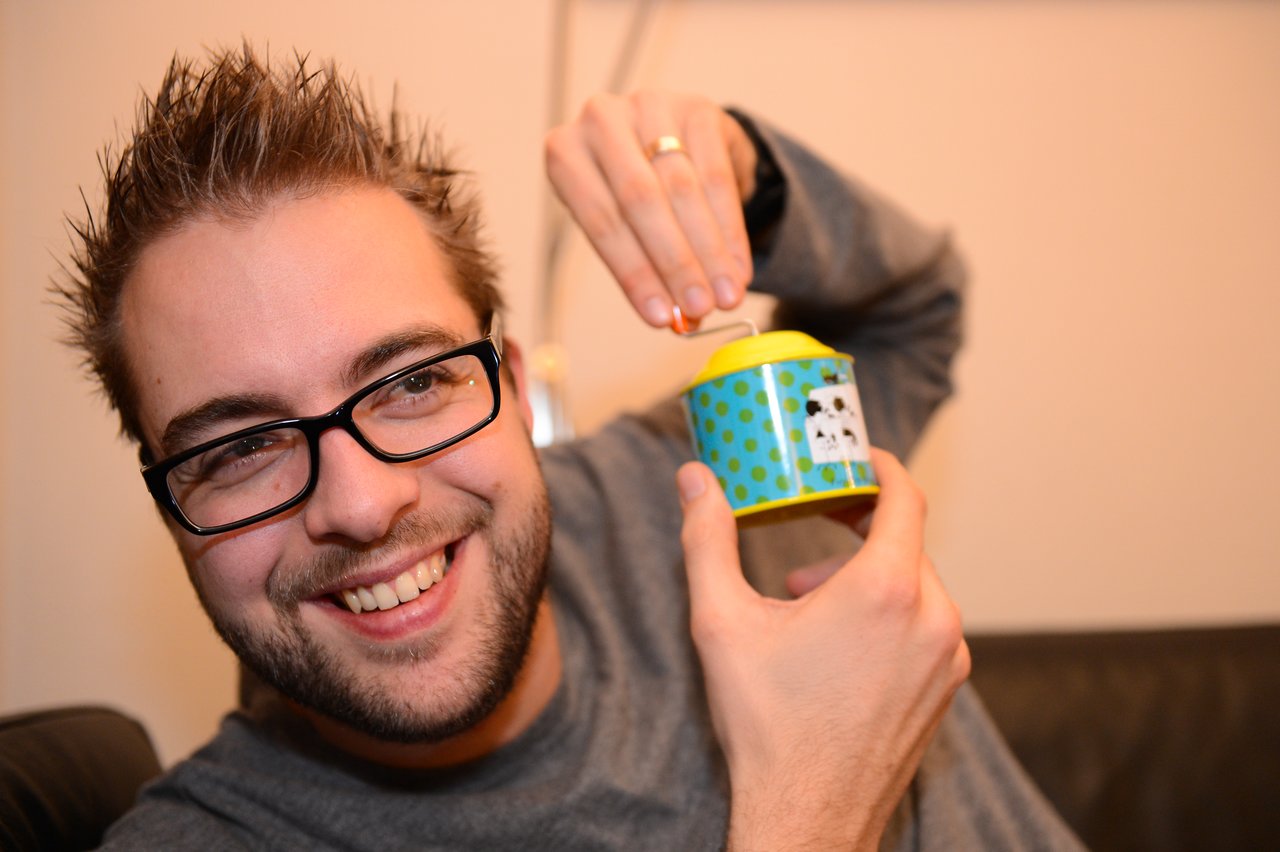 A smiling man with glasses turns the handle of a small, colorful music box at a party.