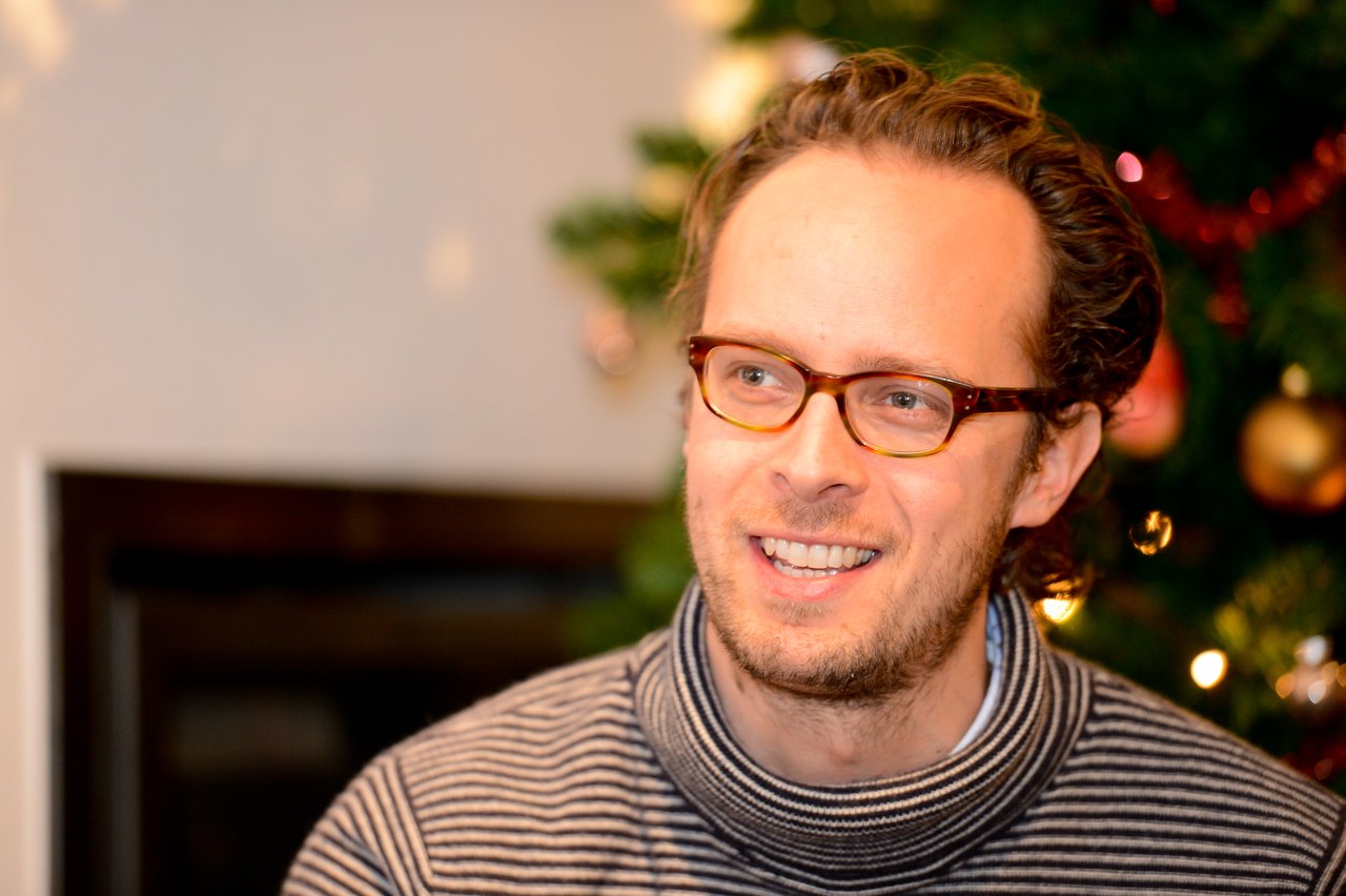 A man wearing glasses and a striped sweater smiles in front of a decorated Christmas tree at a party.