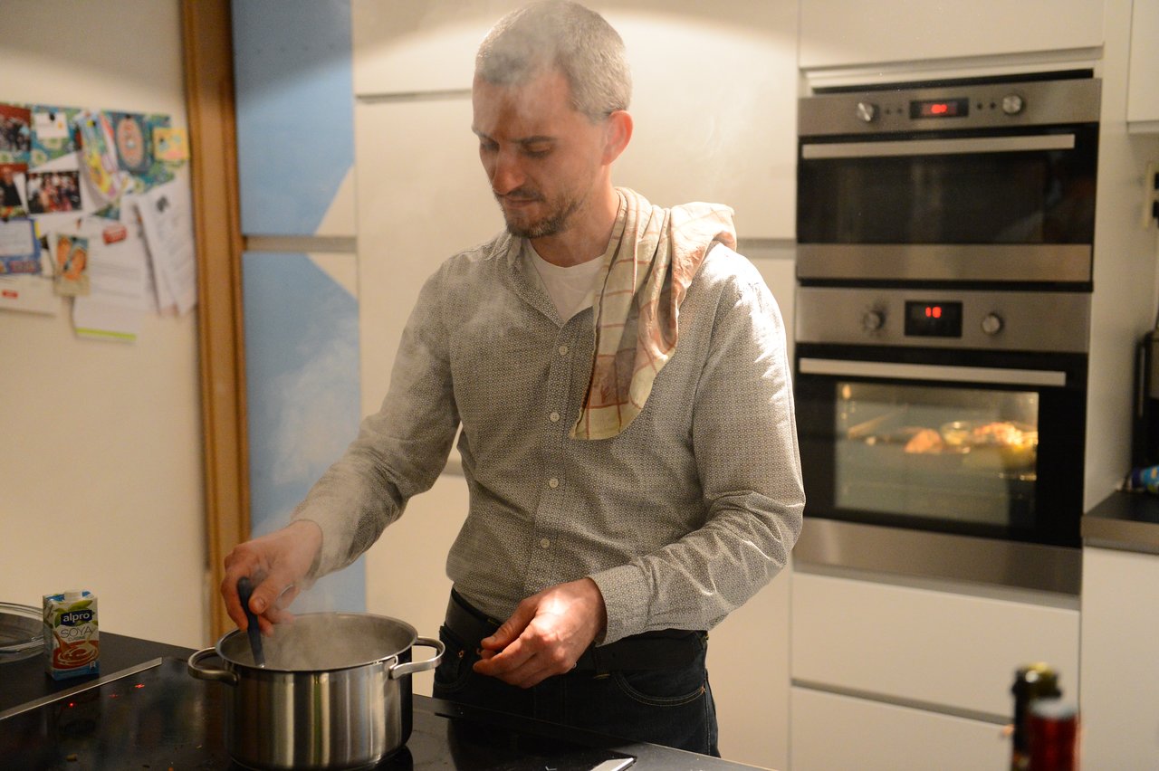 A man stirs a steaming pot on the stove while cooking in a kitchen, with food baking in the oven.