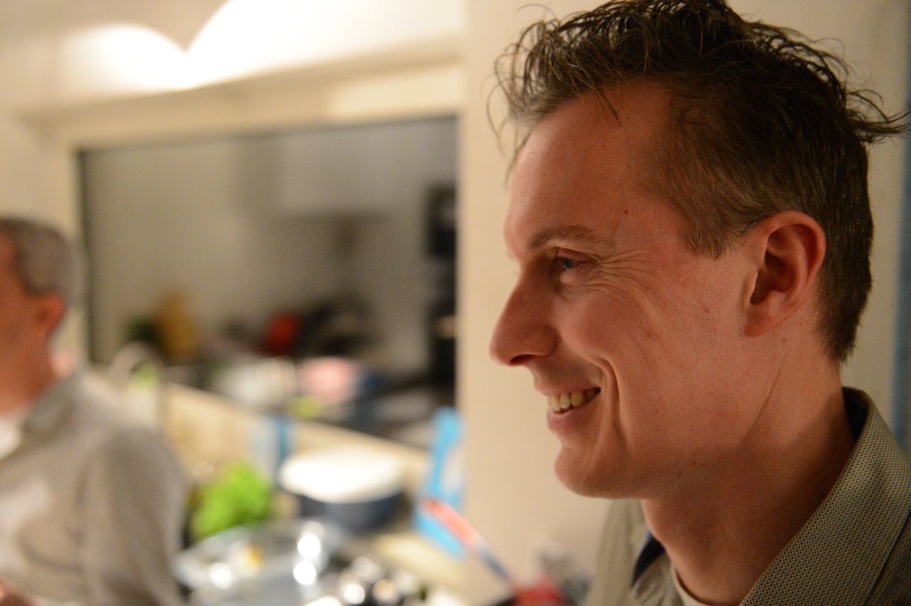A man smiles while enjoying a New Year's Eve party, with another guest and kitchen items in the background.