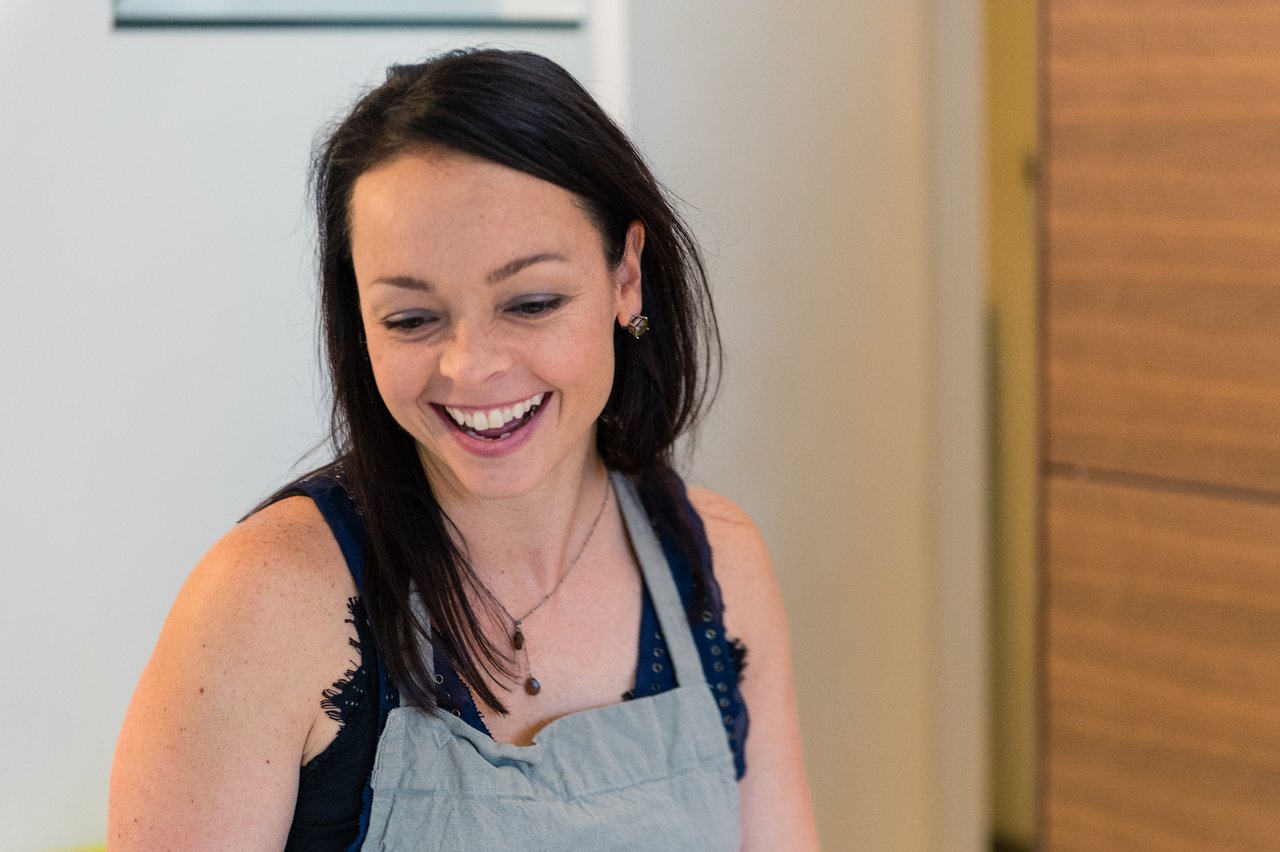 A woman with dark hair and a gray apron smiles while looking down at something off-camera.